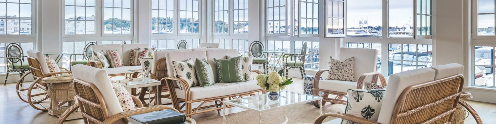 A sunlit room with large windows with a view of the harbor, wicker and wood furniture with decorative pillows, and a coffee table.