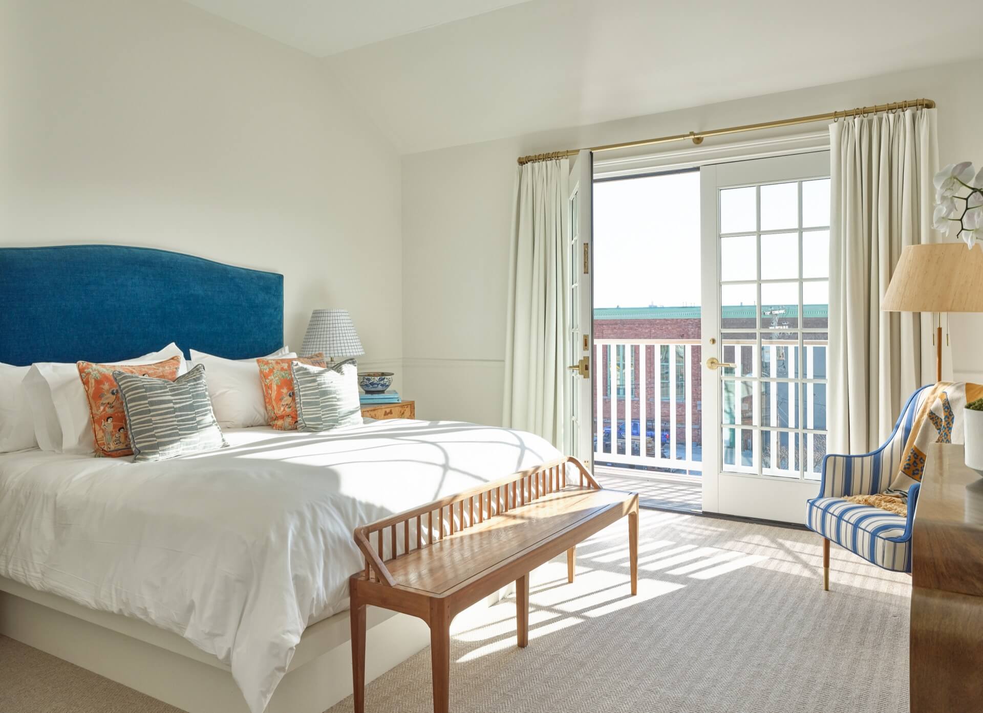 A hotel room with a large bed, balcony door, wooden bench, and striped chair.