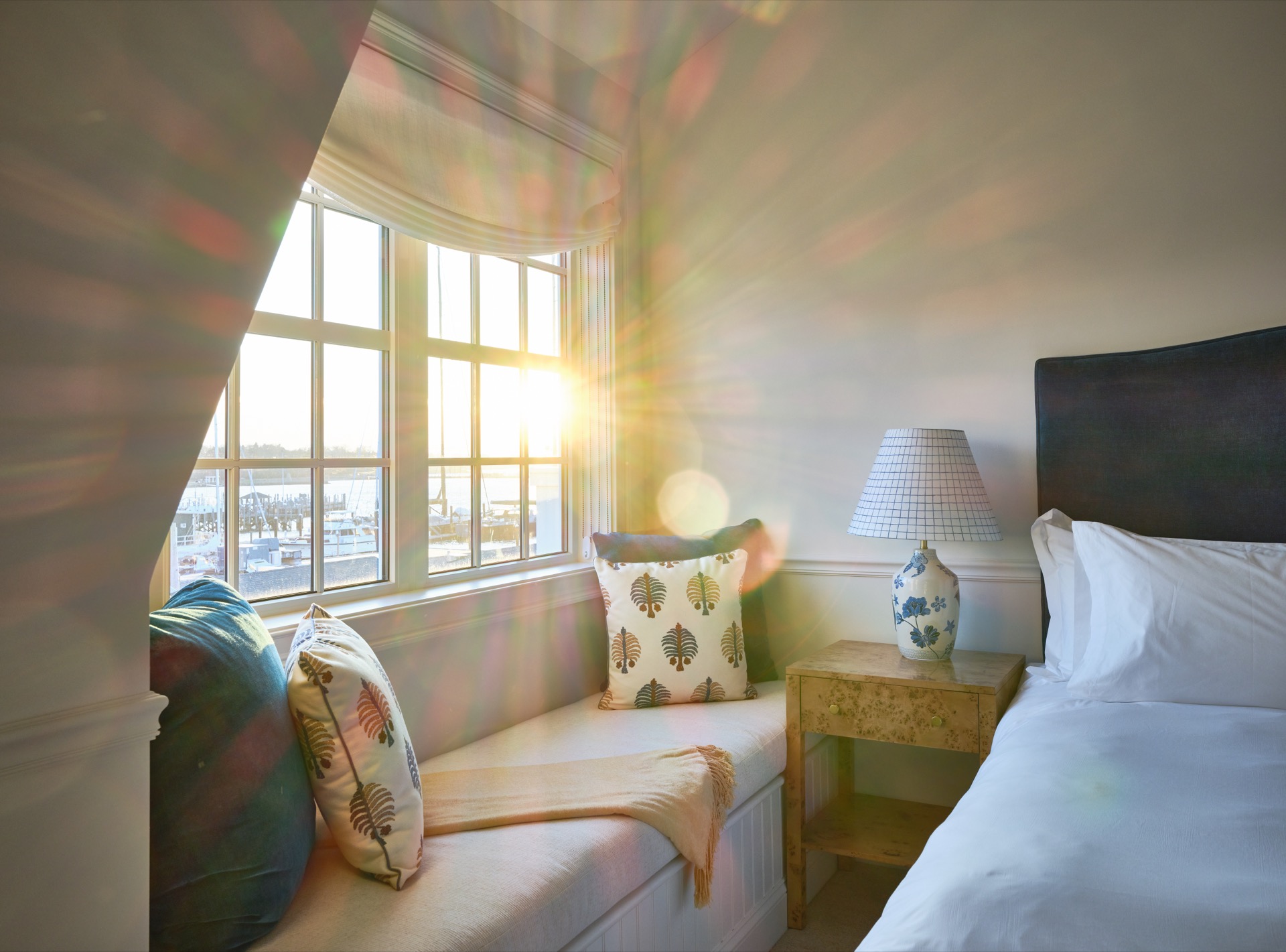 A bright and cozy hotel room with a window seat, a bed, and a lamp.