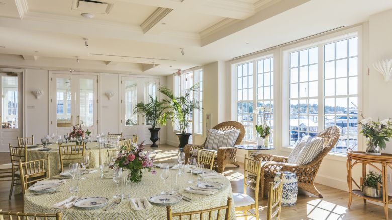 A bright event venue with round tables, chairs, elegant table settings, floral centerpieces, and large windows with harbor views.