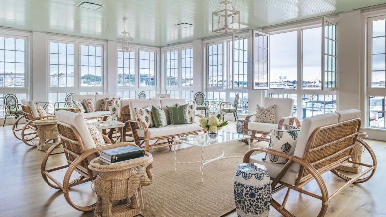 A sunlit room with large windows with a view of the harbor, wicker and wood furniture with decorative pillows, and a coffee table.