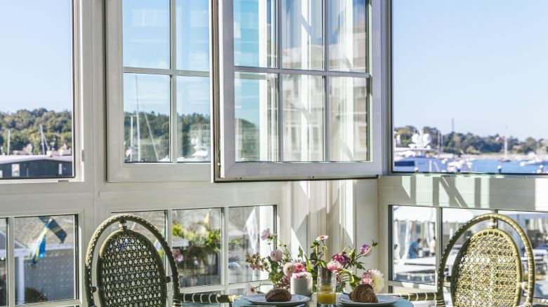 A sunlit room featuring a small table set with food, two chairs, and a large window with a scenic harbor view outside.