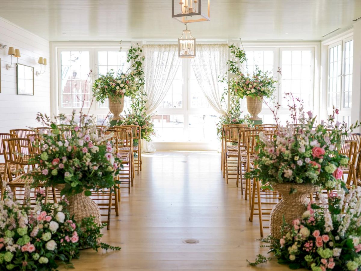 An elegantly decorated indoor RI wedding venue with floral arrangements, wooden chairs lining the aisle, and large bright windows.