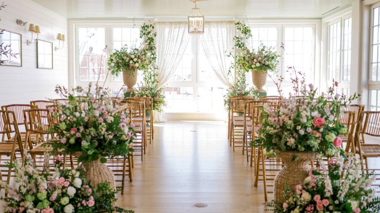 An elegantly decorated indoor RI wedding venue with floral arrangements, wooden chairs lining the aisle, and large bright windows.