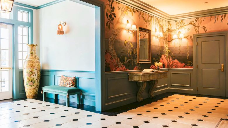 The front hall featuring elegant wallpaper, a green bench with cushions, a large vase, a mirror above a table, and soft lighting.