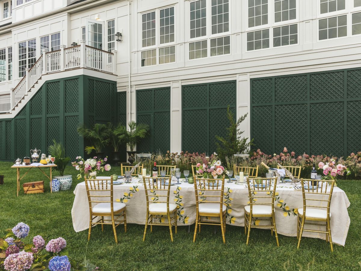 An outdoor setting features a long table with floral decorations and gold chairs, set against a white building with a staircase.