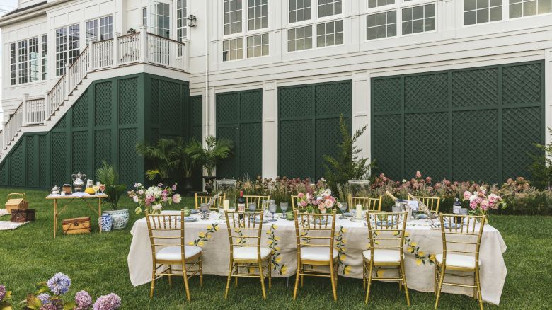 An outdoor setting features a long table with floral decorations and gold chairs, set against a white building with a staircase.