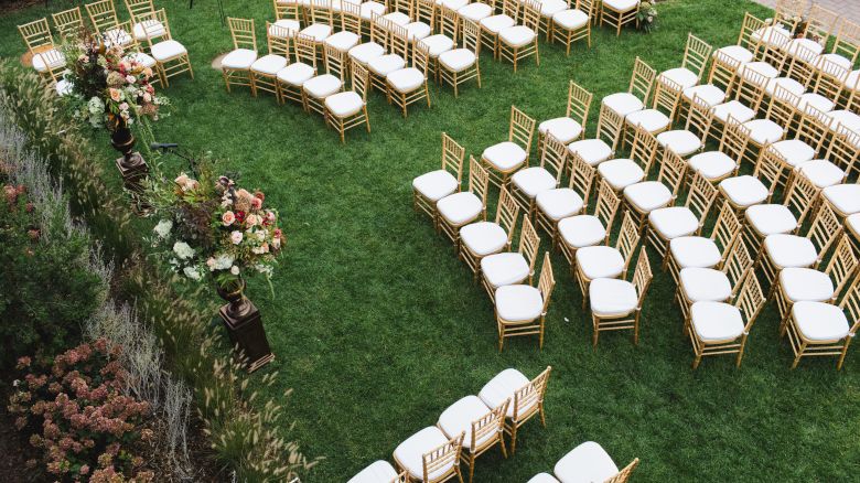 An outdoor event setup with rows of white-cushioned chairs arranged on the lawn surrounded by flowers and greenery.