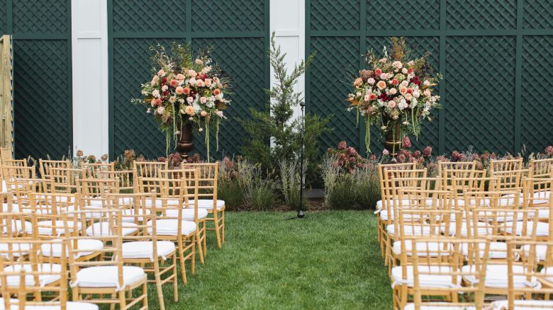 An outdoor wedding ceremony setup with rows of chairs and two large flower arrangements at the front, and a lattice backdrop.