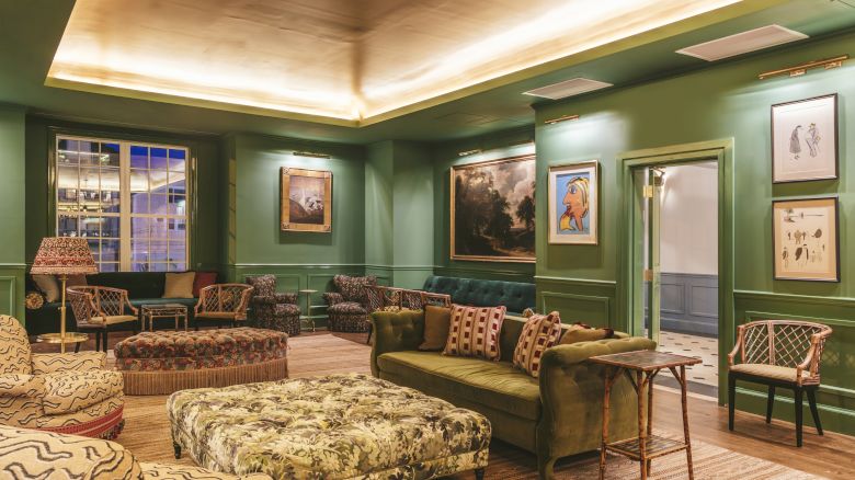 The Studio Bar decorated with green walls, wicker chairs, a plush coffee table, a green couch, framed art, and warm lighting.
