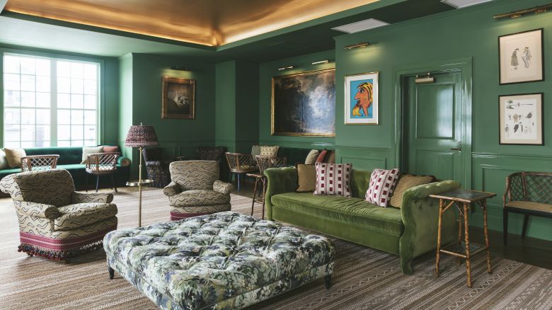The Studio Bar decorated with green walls, artwork, booth seating, a large window, round tables, and wicker chairs.
