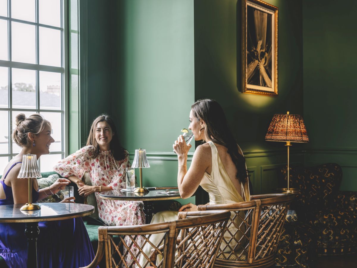 Three women sitting in a stylish, green-painted room with a large window, enjoying drinks and conversation at a small table.