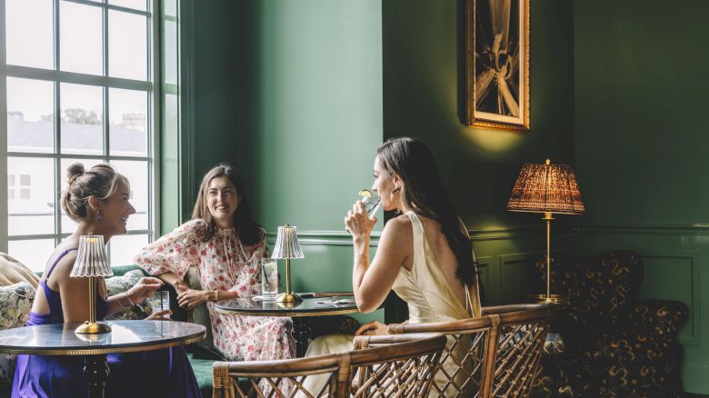 Three women sitting in a stylish, green-painted room with a large window, enjoying drinks and conversation at a small table.