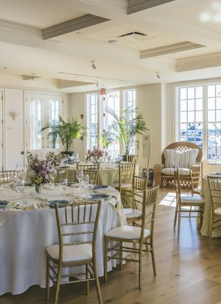 A bright event venue with round tables, chairs, elegant table settings, floral centerpieces, and large windows with harbor views.