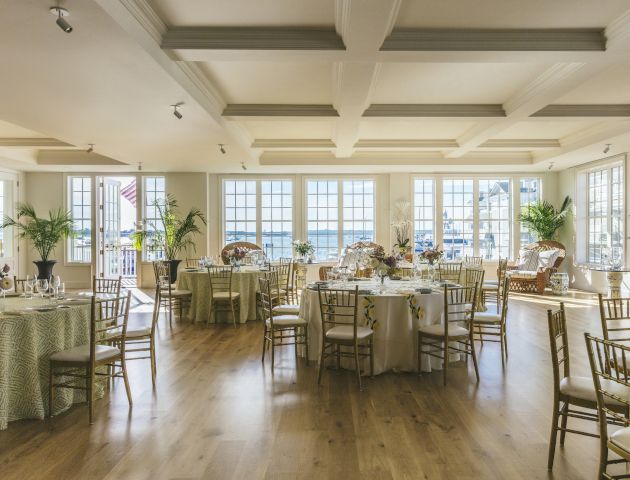 A brightly lit room with tables and chairs arranged for an event, featuring large windows, wooden floors, and elegant decor.
