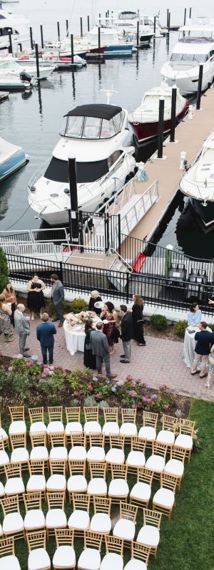 A waterfront wedding setup with rows of chairs on a lawn, guests gathered on a patio, and several boats docked at a marina.