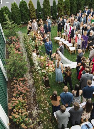 A wedding ceremony taking place outdoors with guests standing watching the bride and groom up in front of a floral display.