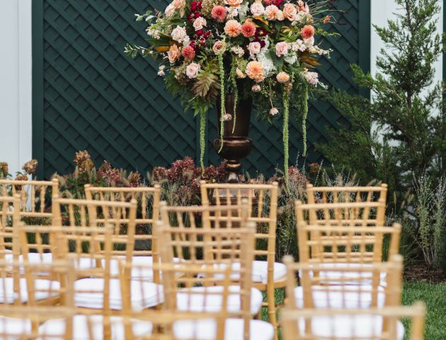 Rows of wooden chairs face a large floral arrangement in an outdoor setting, likely prepared for a wedding or formal event.