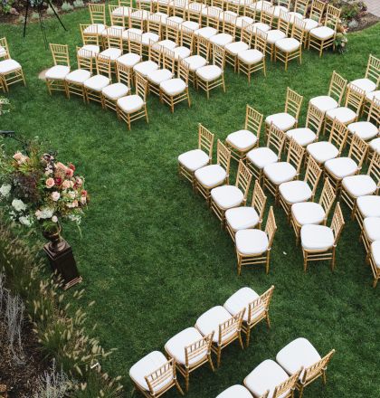 An outdoor event setup with rows of white-cushioned chairs arranged on the lawn surrounded by flowers and greenery.